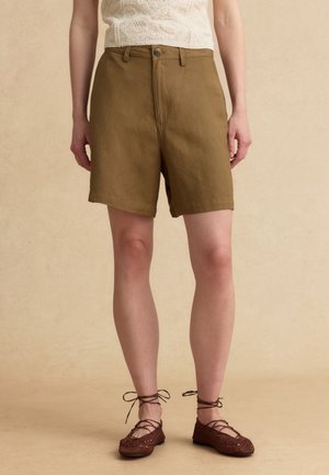 Vrouwelijk model draagt kaki shorts met hoge taille, crème gebreide mouwloze top en bruine gevlochten veterschoenen, staand op een beige oppervlak.