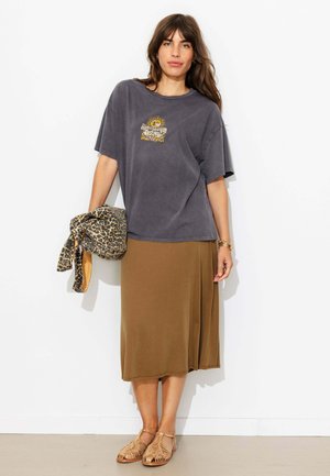 Femme portant un t-shirt ample gris foncé avec motif graphique, jupe midi marron, sandales tissées beige, tenant une veste à imprimé léopard sur fond blanc.