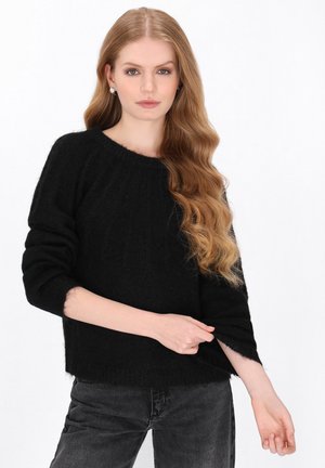 Giovane donna con lunghi capelli biondi ondulati che indossa un maglione nero peloso e jeans scuri su uno sfondo bianco uniforme.