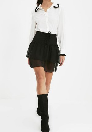 Frau trägt ein weißes Hemd mit schwarzen Schleifendetails, einen schwarzen, geschichteten Minirock und schwarze kniehohe Stiefel und geht nach vorne.