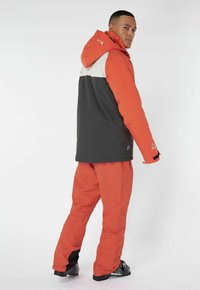 Orange und schwarze Schneejacke mit Kapuze, die ein kontrastierendes graues Paneel aufweist. Weicher Stoff und verstellbare Bündchen. Kombiniert mit passenden Hosen.