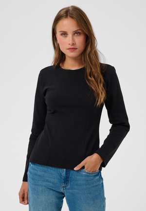 Jeune femme aux longs cheveux bruns portant un haut noir à manches longues et un jean bleu, debout avec une main dans la poche.
