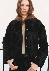 Giacca di velluto nero con colletto in pile marrone, dotata di tasche frontali e bottoni argentati, indossata sopra un top crema.
