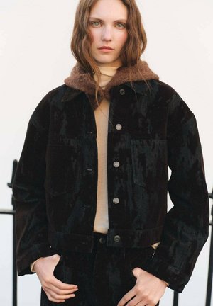 Chaqueta de pana negra con un cuello de forro polar marrón, con bolsillos frontales y botones plateados, usada sobre una camiseta color crema.