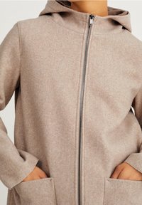 Abrigo con capucha de color beige, con cremallera y hecho de un tejido suave y texturizado. Presenta bolsillos laterales y un cierre de cremallera limpio. Diseño simple y funcional.