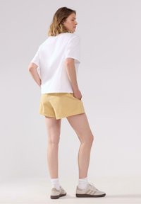 Vit kortärmad t-shirt i kombination med gula rutiga shorts. Modell som bär beige sneakers och vita strumpor, med händerna i fickorna. Enkel design.