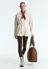 Vrouw draagt een beige jas met ceintuur, bruine legging, beige sokken en sneakers, bruine sjaal, en draagt een grote bruine draagtas.