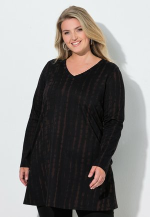 Lachende vrouw met lang blond haar draagt oorringen en een zwarte tuniek met lange mouwen en een V-hals met subtiele verticale patronen.