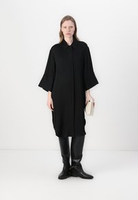 Čierne šaty s dlhým rukávom a golierom, so širokými rukávmi, vyrobené z hladkej látky. Kombinované s elegantnými čiernymi čižmami po kolená.