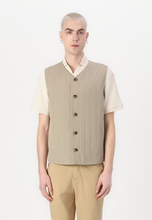 Khaki-Weste mit gestepptem Muster und fünf dunkelbraunen Knöpfen. Kombiniert mit einem kurzärmligen cremefarbenen Hemd und beigen Hosen.