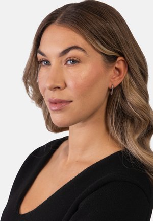 Frau mit welligem hellbraunem Haar, trägt kleine Creolen und schwarzes Oberteil, schaut leicht zur Seite vor einem einfarbigen Hintergrund.