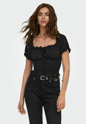 Donna con lunghi capelli ondulati che indossa un top nero arricciato a maniche corte, jeans neri a vita alta, cintura nera e braccialetto argento.