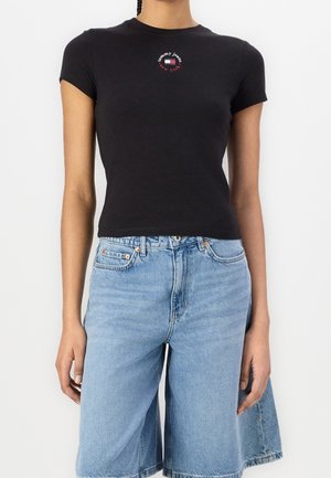 Kvinde iført en tætsiddende sort Tommy Jeans t-shirt og lyseblå denim bermuda-shorts med forlommer og bæltestropper.