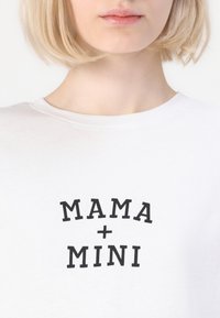 Balta medvilninė marškinėliai su juodu užrašu "MAMA + MINI". Marškinėliai turi klasikį apvalų kaklą ir trumpas rankoves.
