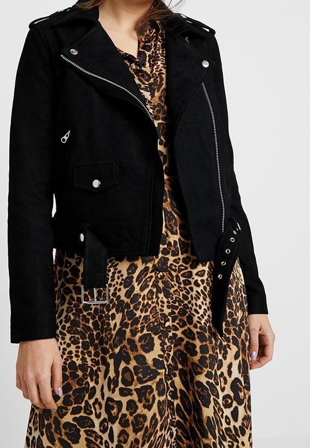 Femme portant une veste de motard noire sur une robe à imprimé léopard marron et noir, debout les bras détendus devant un fond uni.