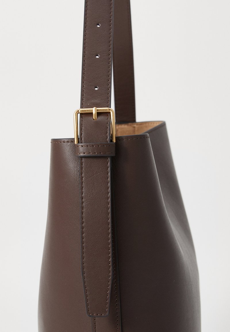 Braune Leder-Schultertasche mit glatter Textur, ausgestattet mit einer goldenen Metallschnalle und einem verstellbaren Tragegurt. Einfaches, nach unten zulaufendes Design.