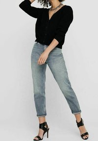 Cardigan nero a costine con scollatura a V, abbinato a jeans tapered azzurri e sandali neri con tacco alto. Una collana a catena arricchisce l'outfit.