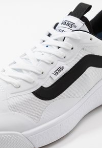 White canvas sneaker featuring a textured surface, black side stripe, thick white sole, and black laces with branding on the tongue.