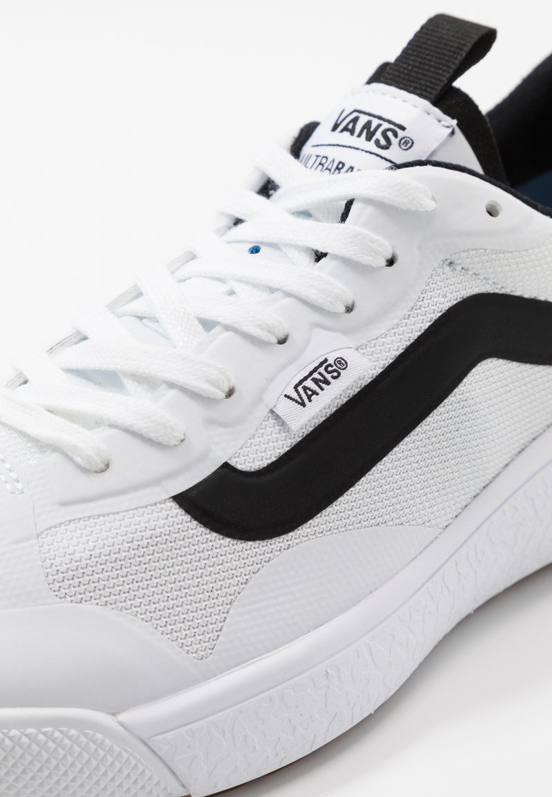 Zapatilla de lona blanca con superficie texturizada, franja lateral negra, suela blanca gruesa y cordones negros con marca en la lengüeta.