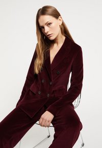 Bordo baršunasti sako s dvostrukim kopčanjem, s reverima, crnim dugmadi i prednjim džepovima, u kombinaciji s odgovarajućim hlačama.