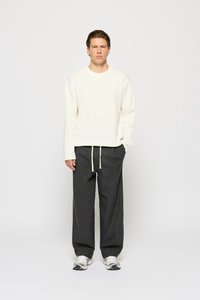 Cream sweater with long sleeves, paired with gray, loose-fitting trousers. White drawstring details and casual sneakers complete the look.