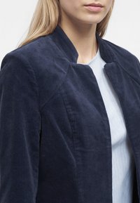 Gros plan d'une femme portant une veste en velours côtelé bleu marine par-dessus un haut bleu clair, montrant le haut du torse et une partie du visage.