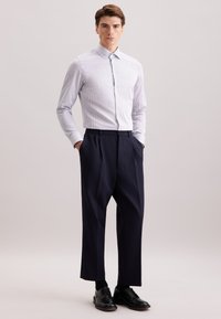 Man staat met zijn handen in zijn zakken, draagt een wit gestreepte langarmshirt, marineblauwe geplooide broek, zwarte sokken en zwarte dress schoenen op een effen achtergrond.