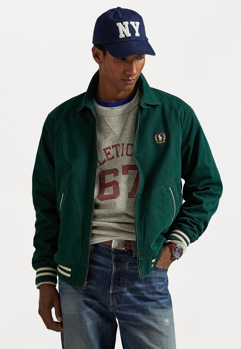 Veste bomber verte avec des rayures blanches sur les poignets, poches zippées, portée par-dessus un sweat à capuche gris. Casquette marine avec "NY" en lettres crème.