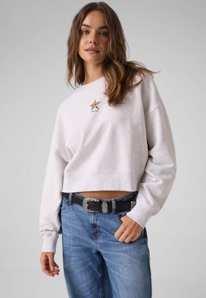 Une femme porte un sweat-shirt blanc court orné d’un petit emblème en forme d’étoile, un jean bleu et une ceinture noire sur un fond gris uni.