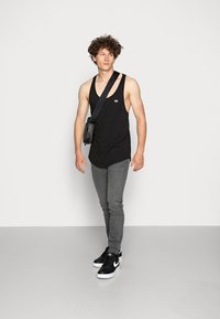 Svart tanktopp, grå slim-fit jeans og svarte sneakers med hvite detaljer. Modellen bærer en liten svart crossbody-veske. Enkelt bakgrunn.