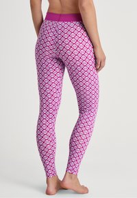 Vrouw die blootsvoets staat in hoge roze leggings met witte geometrische bloempatronen tegen een effen lichte achtergrond.