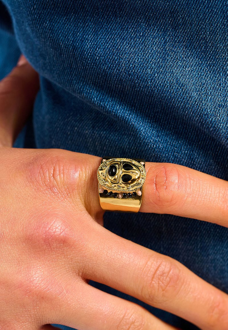 Gold-toned ring with a large, oval black gemstone centerpiece, intricate detailing, and a wide band featuring small decorative stones.