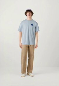 Jauns vīrietis, ģērbies gaiši zila T-kreklā ar tumši zilu ziedu grafiku, bēšās biksēs, baltās sporta kurpēs un gaiši violetā cepurē, stāv pret vienkāršu fona.