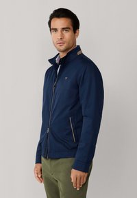 Mężczyzna w granatowej kurtce na zamek z wysokim kołnierzem i oliwkowozielonych spodniach, stojący na tle jednolitego, jasnoszarego tła.