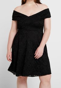 Svart off-shoulder-klänning med texturerat spetsmaterial, en figursydd bodice och en utsvängd kjol med en scallopad nederkant.