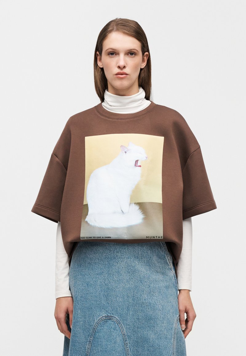 Bruine oversized sweatshirt met een kattenafbeelding, voorzien van een ronde hals en korte mouwen. Draagt over een witte coltrui en een spijkerrok.