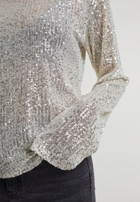 Top argenté à sequins à manches longues porté avec un denim noir, montrant un scintillement texturé et un détail de manche ajustée sur un fond neutre.