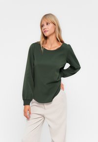 Blouse à manches longues vert foncé avec un col arrondi et des poignets smockés. Matière douce au toucher lisse, avec un ourlet arrondi.