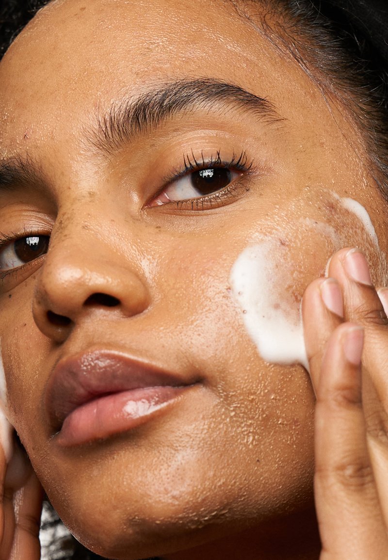 Gros plan sur un visage avec un nettoyant moussant léger appliqué. La peau est bronzée avec une texture visible et de petites imperfections, et les lèvres sont d'un rose doux.