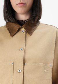 Kvinne i en beige jakke med brun lærkrage og sølvknapper, over en mørk leopardmønstret topp, vist fra haken til brystet.