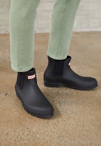 Black waterproof ankle boots with elastic side panels and a textured sole, displaying a red and white "HUNTER" logo on the front.