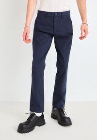 Pantalons ajustés bleu marine à la texture lisse et à la coupe fuselée, accompagnés de chaussures noires à lacets massives avec une semelle épaisse.
