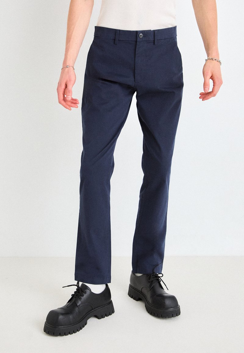 Pantalons ajustés bleu marine à la texture lisse et à la coupe fuselée, accompagnés de chaussures noires à lacets massives avec une semelle épaisse.
