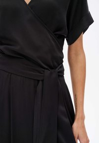 Schwarzes Kleid mit Wickeldesign, kurzen Ärmeln und gebundenem Taillenbund. Der glatte Stoff hat einen leicht schimmernden Effekt. Keine sichtbaren Muster oder Hardware.