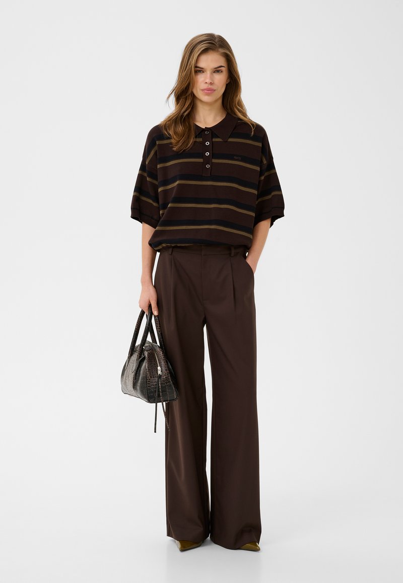 Femme debout portant un polo rayé marron, un pantalon marron à jambes larges, tenant un sac à main texturé foncé devant un fond gris uni.