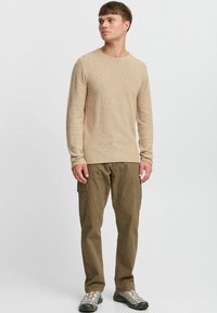 Beige langärmliger Strickpullover mit Rundhalsausschnitt, kombiniert mit olivgrünen Cargohosen mit Seitentaschen und grauen Sportschuhen.