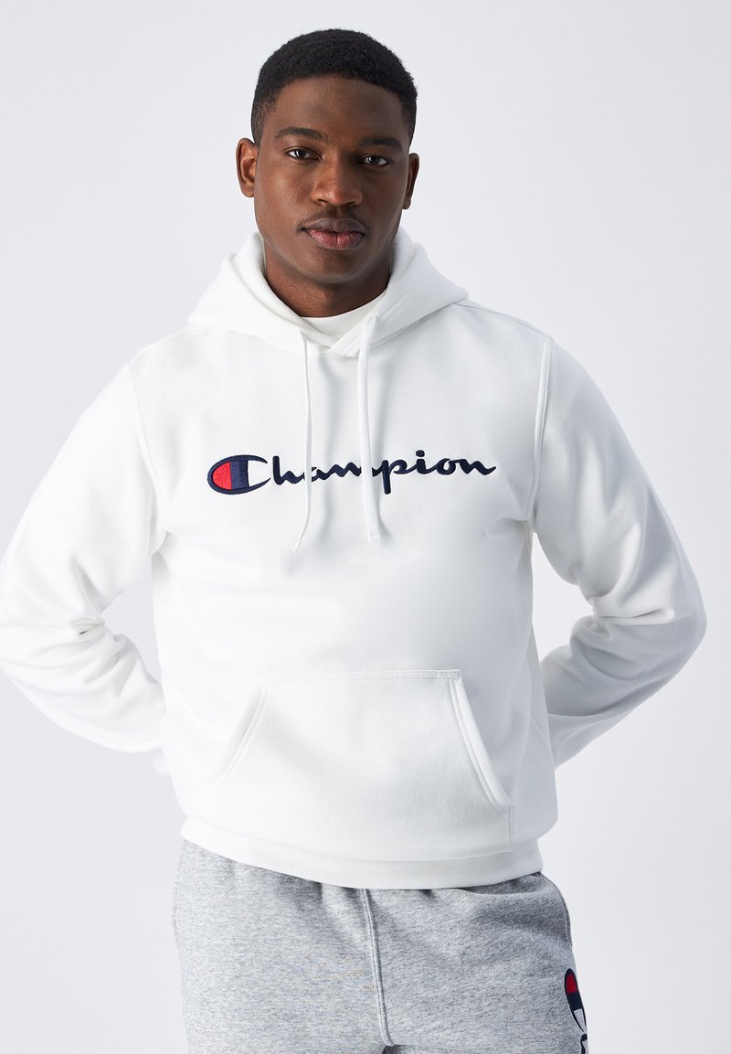 Biela mikina s kapucňou a prednou kapsou, s námorníckym a červeným logom "Champion". Tkanina vyzerá jemne a štruktúrovane. Kombinovaná so sivými teplákmi.