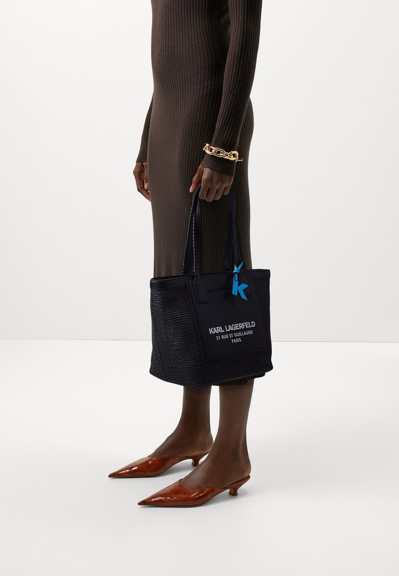 Black woven tote bag with blue accents, featuring the text “KARL LAGERFELD” and a charm. Worn with a brown ribbed dress and pointed shoes.