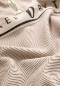 Beige strukturierter Stoff mit einem gerippten Muster, ausgestattet mit einem schwarzen Logo-Band und einem metallischen Reißverschluss. Das Material wirkt weich und langlebig.