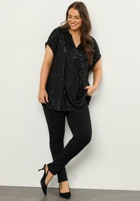 Schwarzes Paillettenoberteil mit V-Ausschnitt, kurzen Ärmeln und lockerer Passform, kombiniert mit schwarzen, eng anliegenden Hosen und schwarzen High Heels.